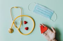 Top view of crop anonymous person hand with red paper heart on table with stethoscope and medical mask for coronavirus prevention